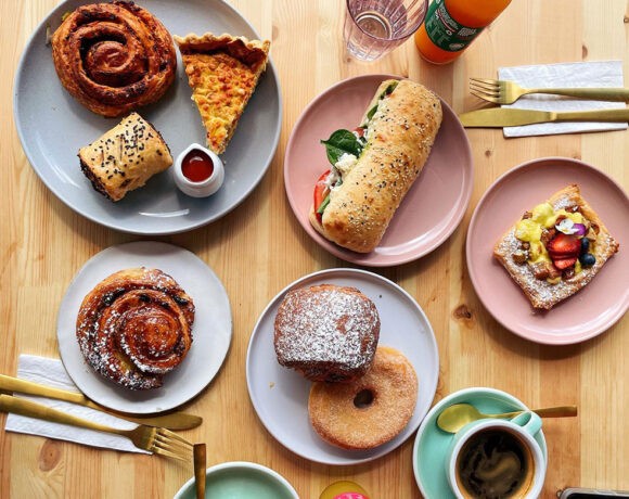 a wooden table with a brunch spread on different pastel plates, there are various sweet and savoury baked goods and a cup of coffee as well as cutlery (miss sina)