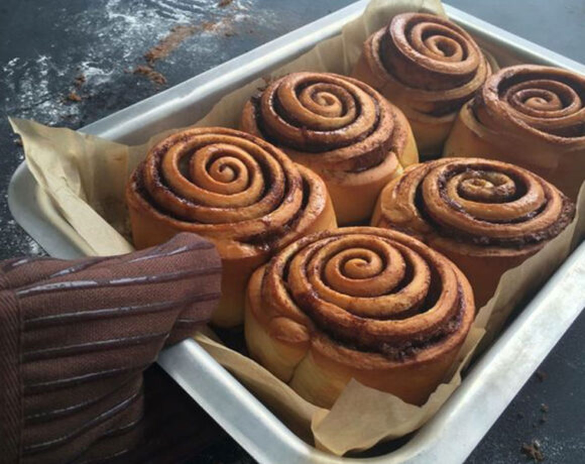 A hand wearing an oven glove places a baking tray full of cinnamon rolls onto a countertop (Cinnabon)