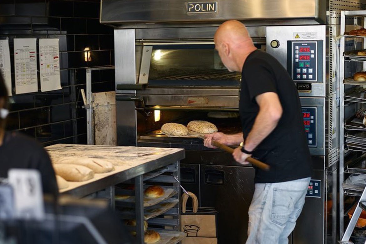 A baker pulls loaves of bread out of a giant bread oven on bread pallets (The Grumpy Baker)