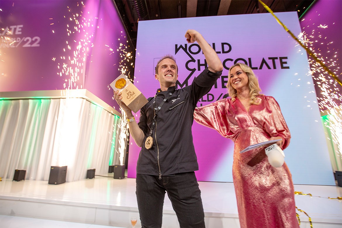 Winner of the World Chocolate Masters, Lluc Crusellas, stands on a stage holding aloft his trophy. A woman in a pink dress stands next to him.
