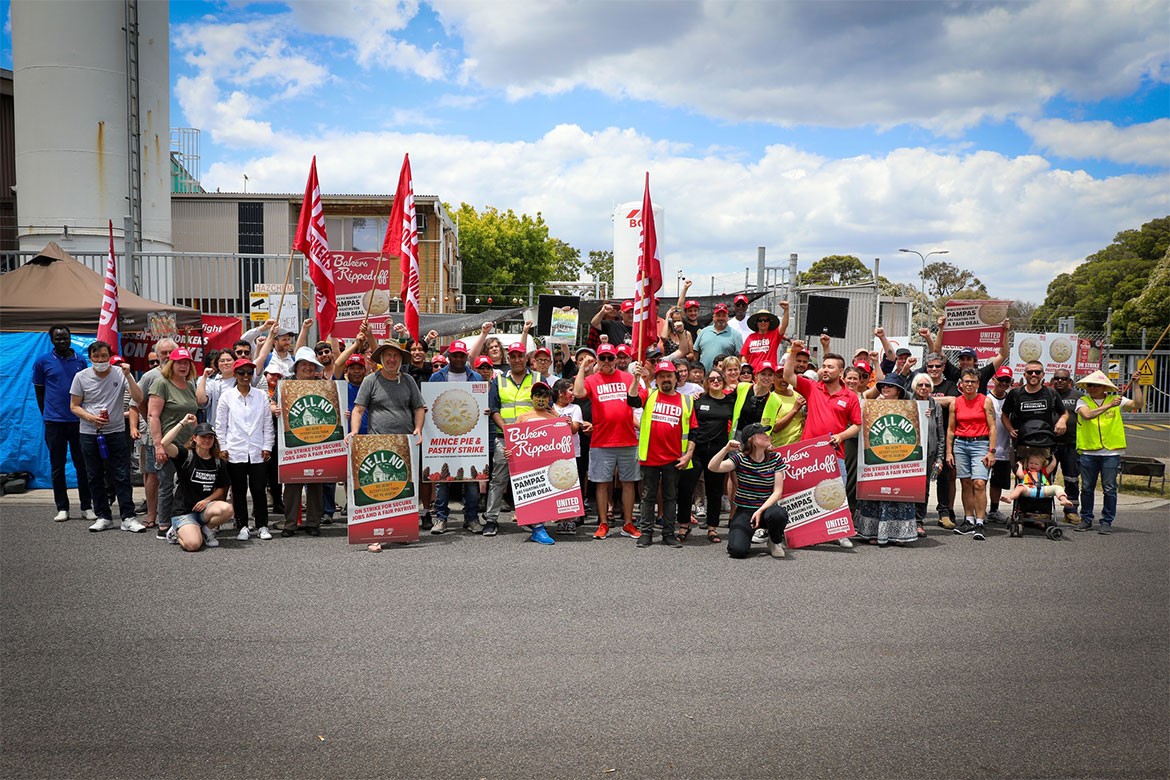 A group of striking Pampas Pastry workers stands in front of a factory holding signs and placards in protest