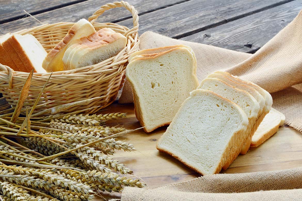 A few slices of white bread sit on a piece of hessian next to a basket with some white bread, there is some wheat there as well (fibre)