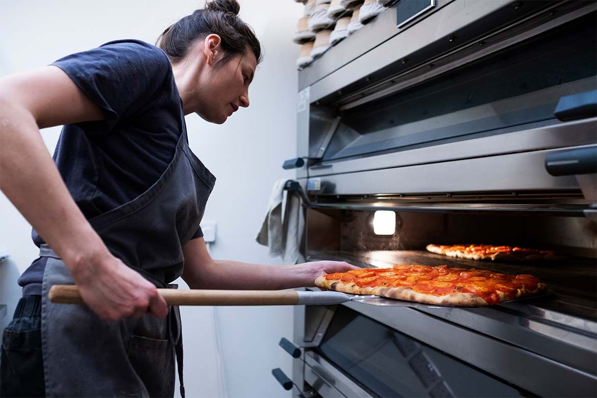 Woman wearing apron standing in an artisan bakery, placing pizza into oven (The People's Oven)