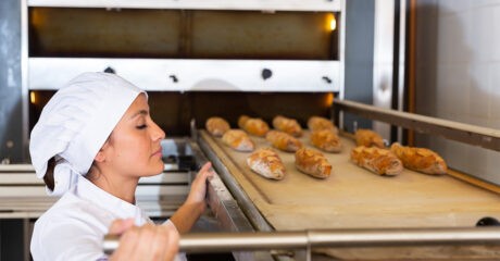 Young Hispanic woman in white uniform working in bakery, pulling freshly baked baguettes out of industrial oven (flexible)