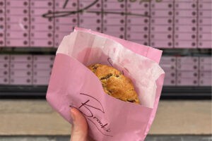 a hand holding a choc chip cookie in a pink Brooki Bakehouse bag in front of the Brooki Bakehouse glass shopfront