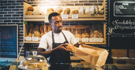 a baker stands behind a bakery counter sliding a loaf of bread into a brown paper bag (industrial relations)