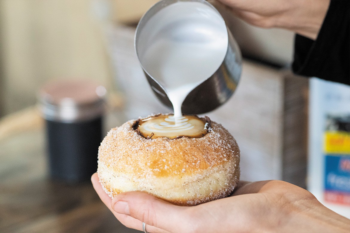 Someone pouring frothed milk into a doughnut with the middle carved out (like a cup) (Kenilworth)