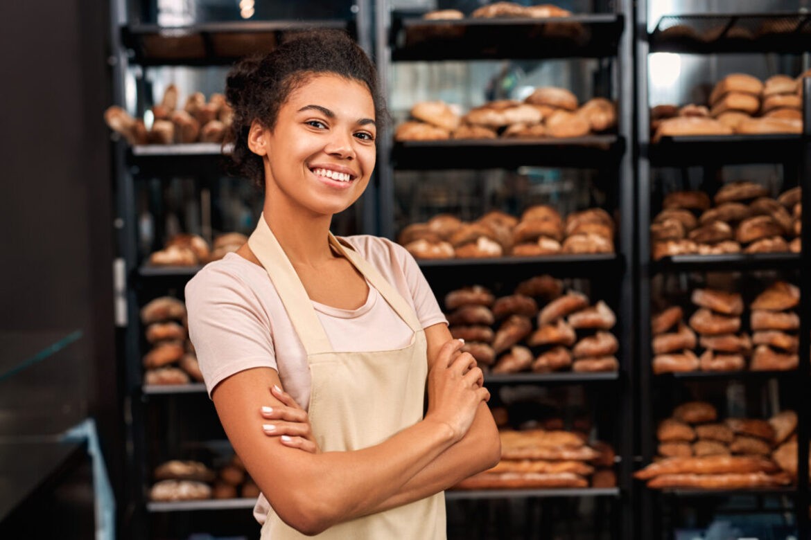 A young lady leans against the doorframe of a bakery. She's smiling