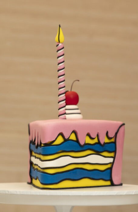 The comic cake, made up of blue and yellow layers with pink icing, is on a plate against a wooden wall.