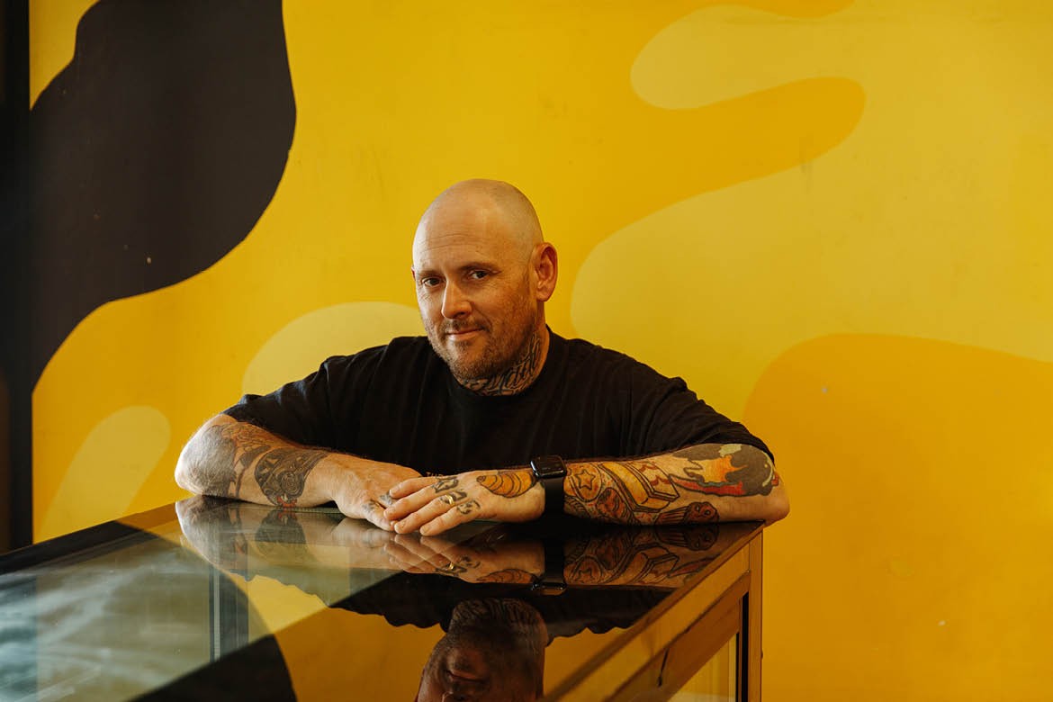 Jason Spencer from Banana Boogie leans on a glass counter in front of a yellow wall.