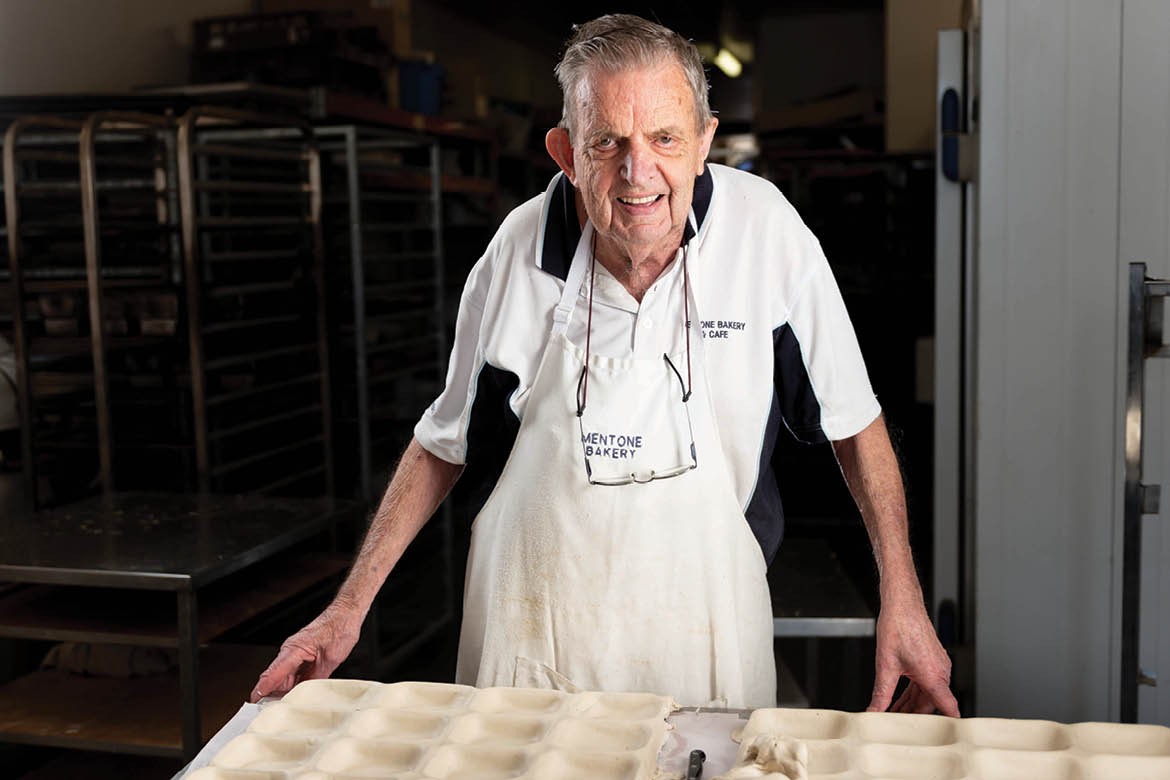 Bill Fuller wears a Mentone Bakery apron, standing behind a workbench. He's smiling at the camera.