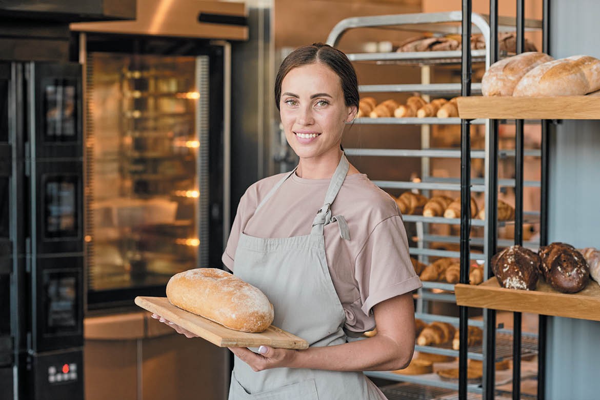 A young woman wearing an apron smiles at the camera while holding a loaf of bread.