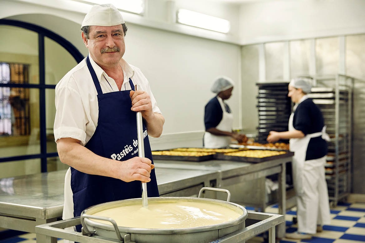 A baker with a grey moustache stirs a large pot of custard. He's looking at the camera.