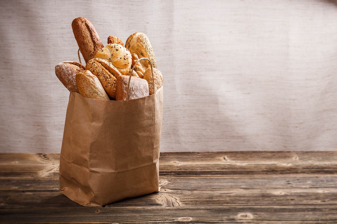 Loaves of bread and baguettes are in a paper bag, that is standing up on a wooden table.