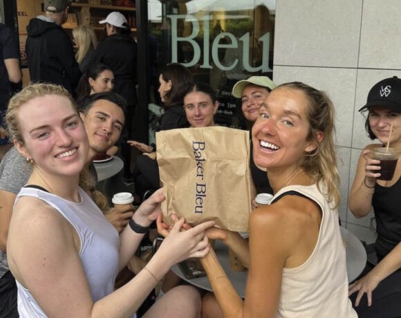 Croissant Run Club runners sit at a table at Baker Bleu, drinking coffees and holding up a paper bag.