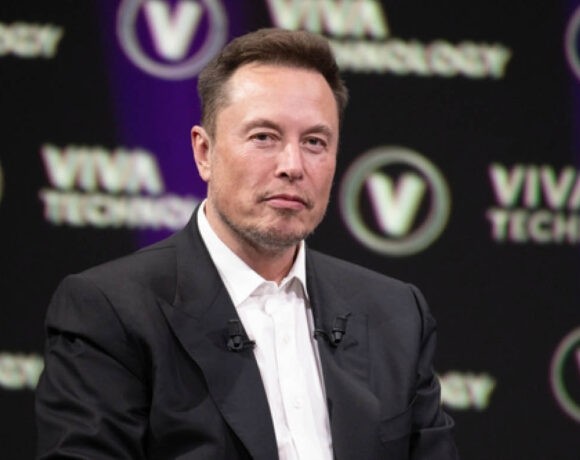 Tesla owner Elon Musk is pictured in a dark suit with a white shirt. He's in front of a dark background.