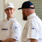 Bjarke Svendsgaard smiles at one of his teachers. He's wearing chef whites and wearing a white cap. His teacher has his back to the camera and is wearing a black cap.