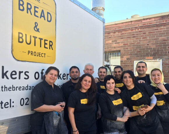 The Bread and Butter Project team poses in front of a white sign that bears the yellow Bread and Butter Project sign. Each team member wears a black shirt.