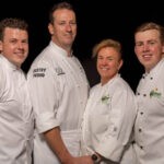 Tracy and Vicki Nickl with their two sons. All four wear chefs whites and are smiling at the camera. There is a black background