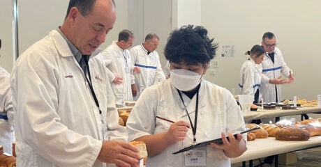 BIEWA competition judges in white coats examine baked good set out in a room full of long tables covered with white tablecloths
